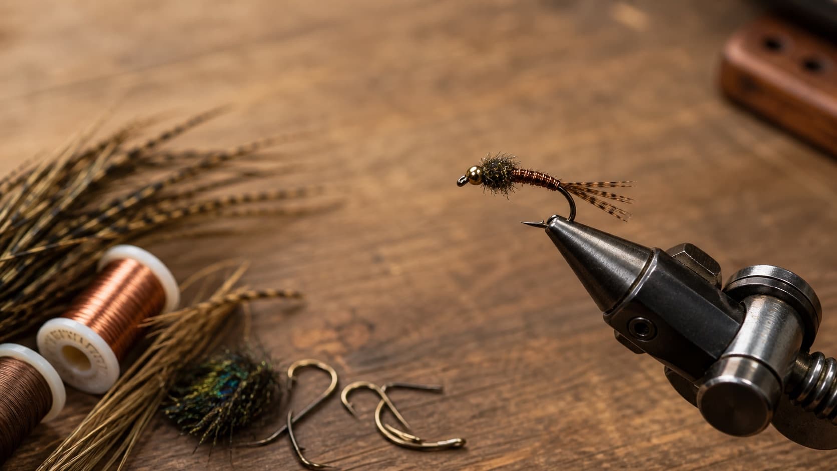 Finished Pheasant Tail Nymph in a fly-tying vise beside copper wire and natural tying materials